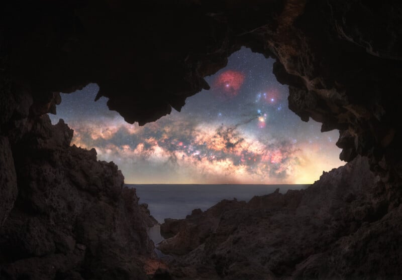 View from inside a rocky cave looking out to a star-filled night sky and the Milky Way over a calm sea, with rugged rocks framing the scene.