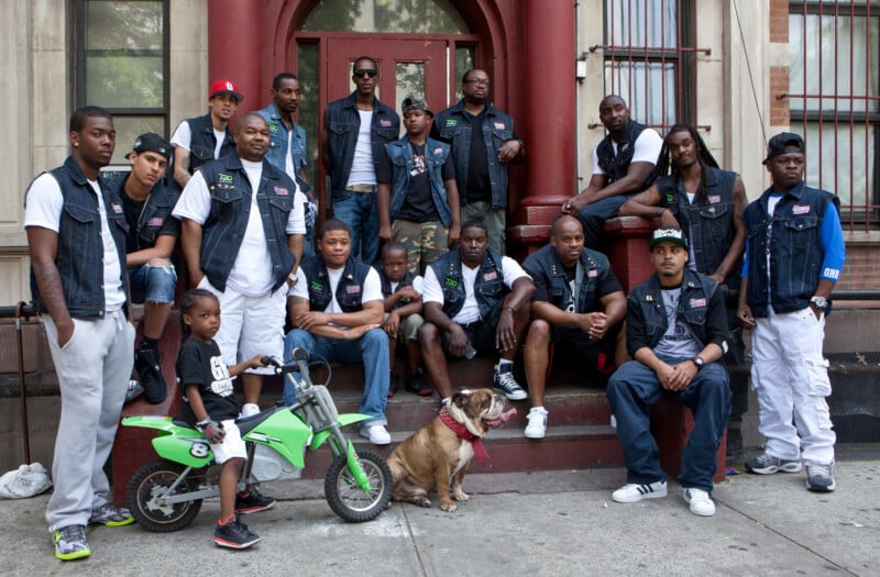 A group of men and boys, some wearing matching sleeveless vests, pose together on the steps of a city building. A bulldog sits in front, and a young boy sits on a small green dirt bike.