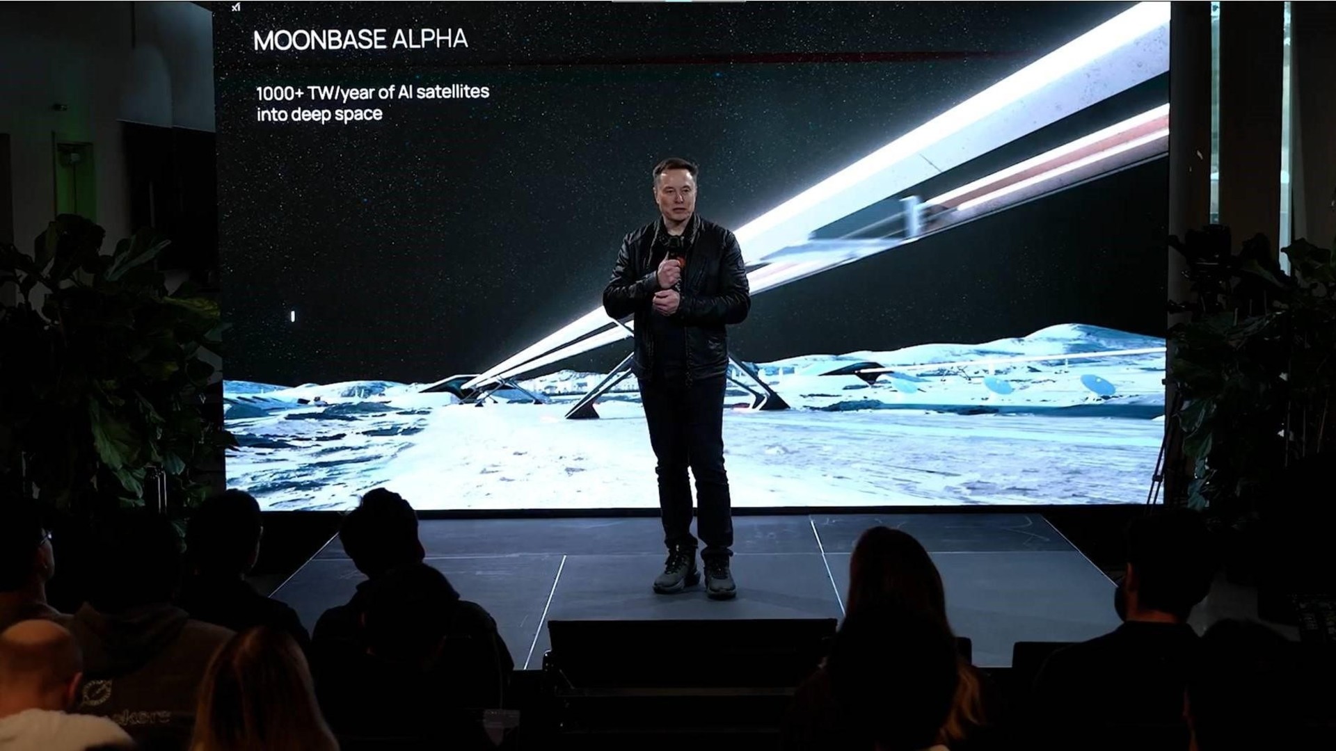a man in a black t-shirt and black jeans speaks into a microphone on a stage in front of an illustration of a railroad-like structure on the moon