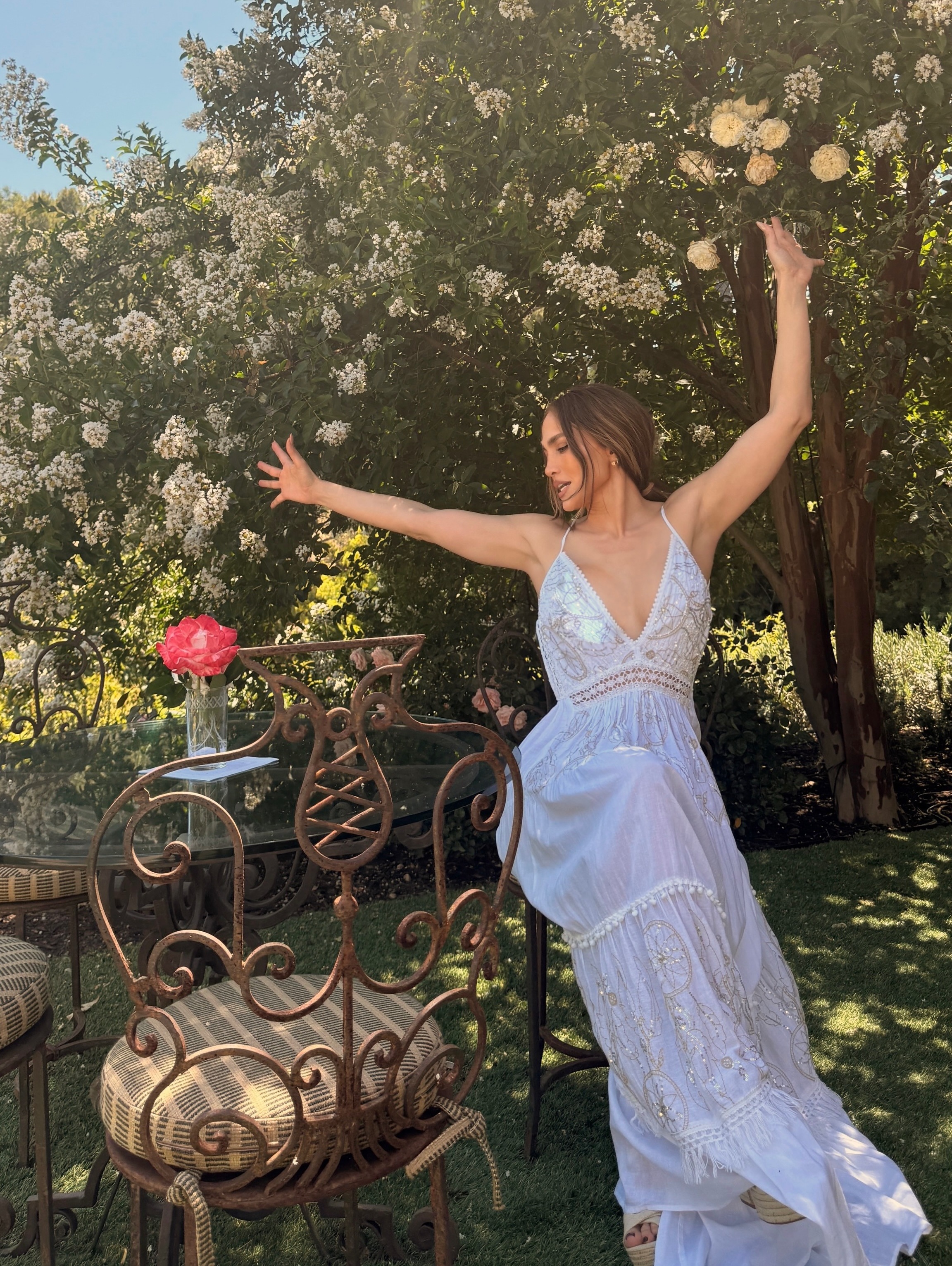 Jennifer Lopez sits outside wearing a white embroidered maxi dress with light brown wicker wedges.
