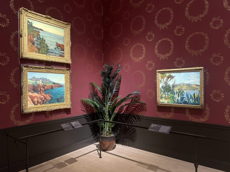 Three framed landscape paintings hang on ornate red wallpaper above a potted plant in a museum gallery, with information plaques displayed below.