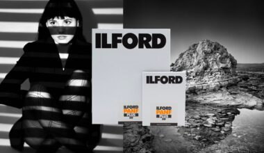 A woman with dramatic light and shadow lines across her face and legs sits next to two boxes labeled "ILFORD PANF PLUS 50." In the background is a black and white rocky landscape with water.