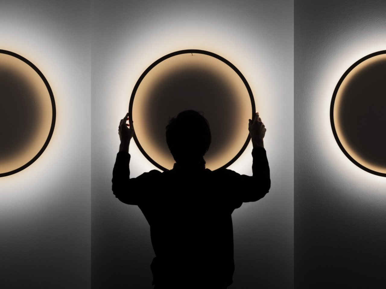 Silhouette of a person standing with arms raised, touching a glowing circular wall light, flanked by two more illuminated circles on a wall.