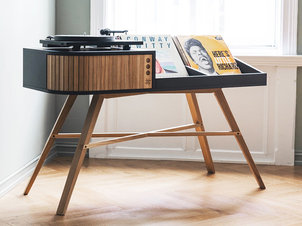 Wooden mid-century record player on a black and wood stand with vinyl records displayed nearby in a bright room.