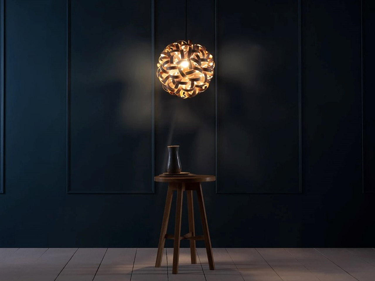 Warm spherical pendant lamp glowing above a wooden stool with a small vase on a round tray in a dark navy room, minimal decor.
