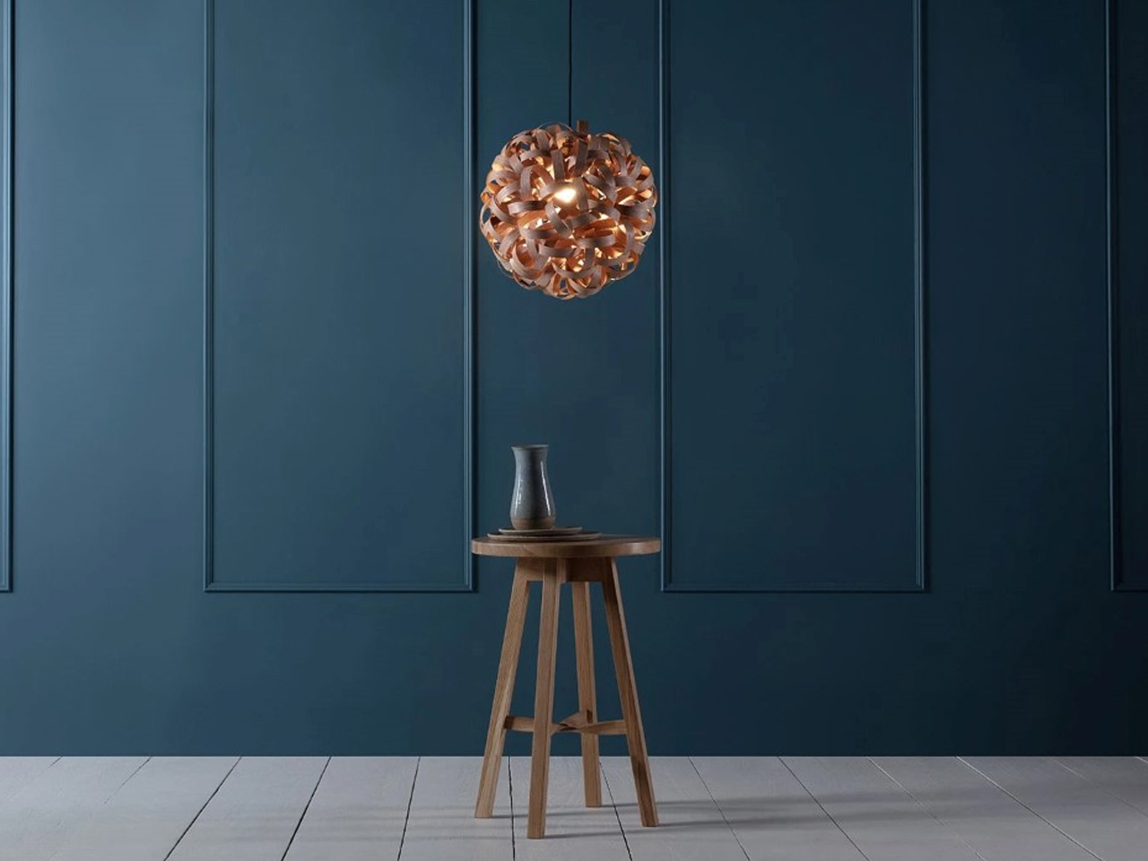 Wooden stool with a small ceramic vase under a copper pendant light against a dark blue paneled wall.