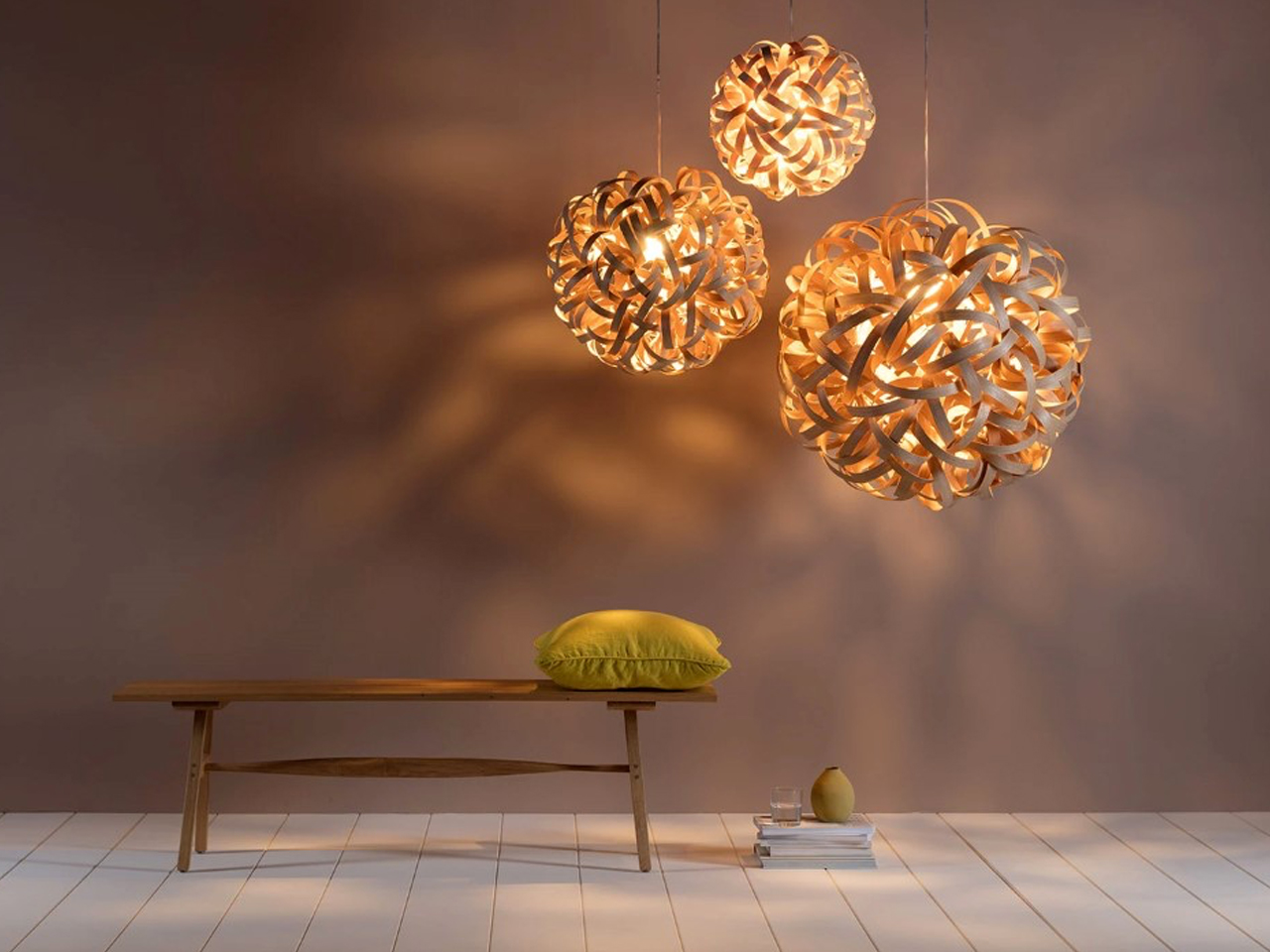 Cozy minimalist room with three warm wooden pendant lights above a wooden bench and a yellow cushion, plus a small coffee setup on the floor nearby.