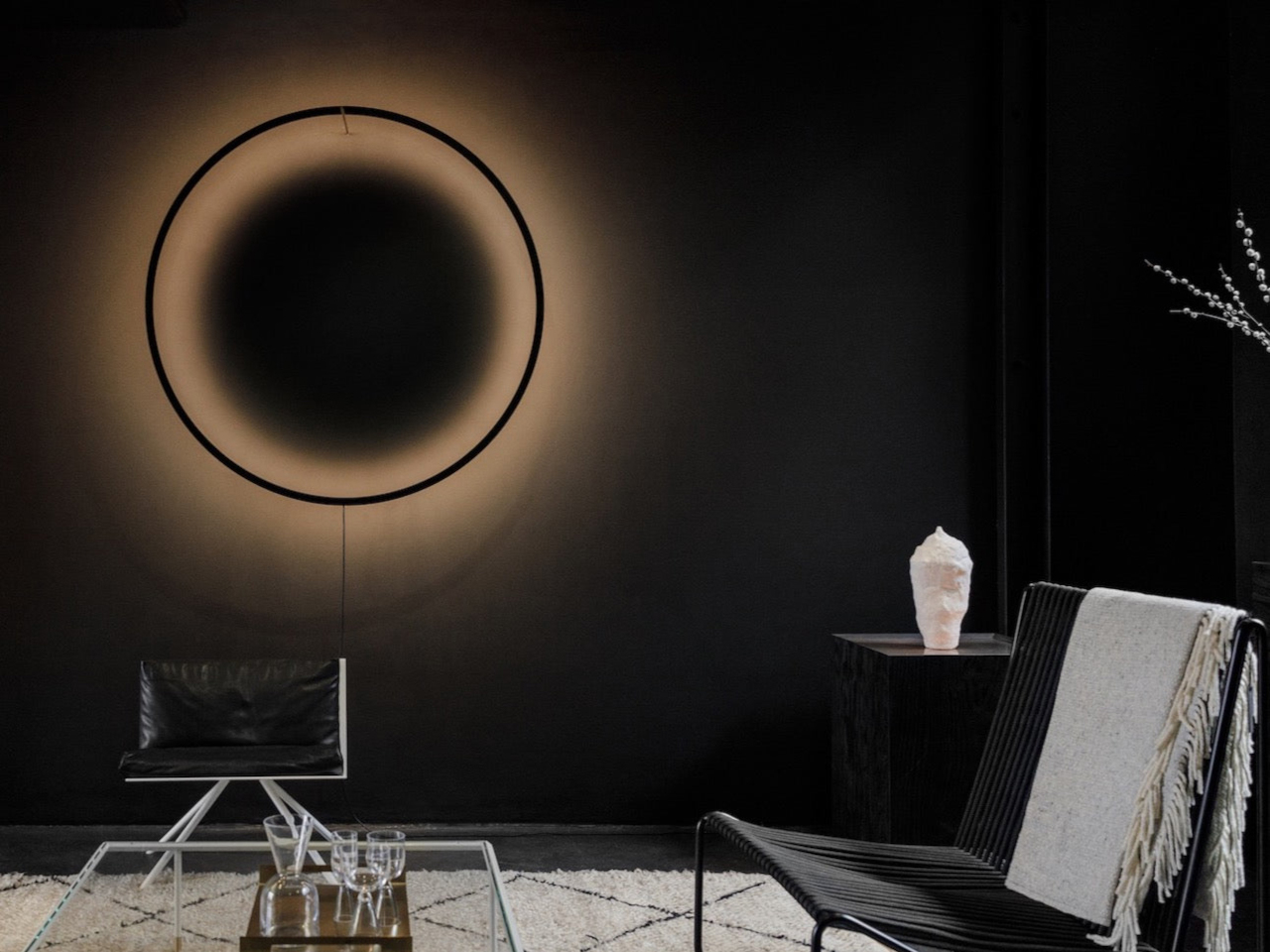 Dark minimalist living room with a large circular wall halo, a black chair, and a glass coffee table; a white sculpture rests on a side table to the right.