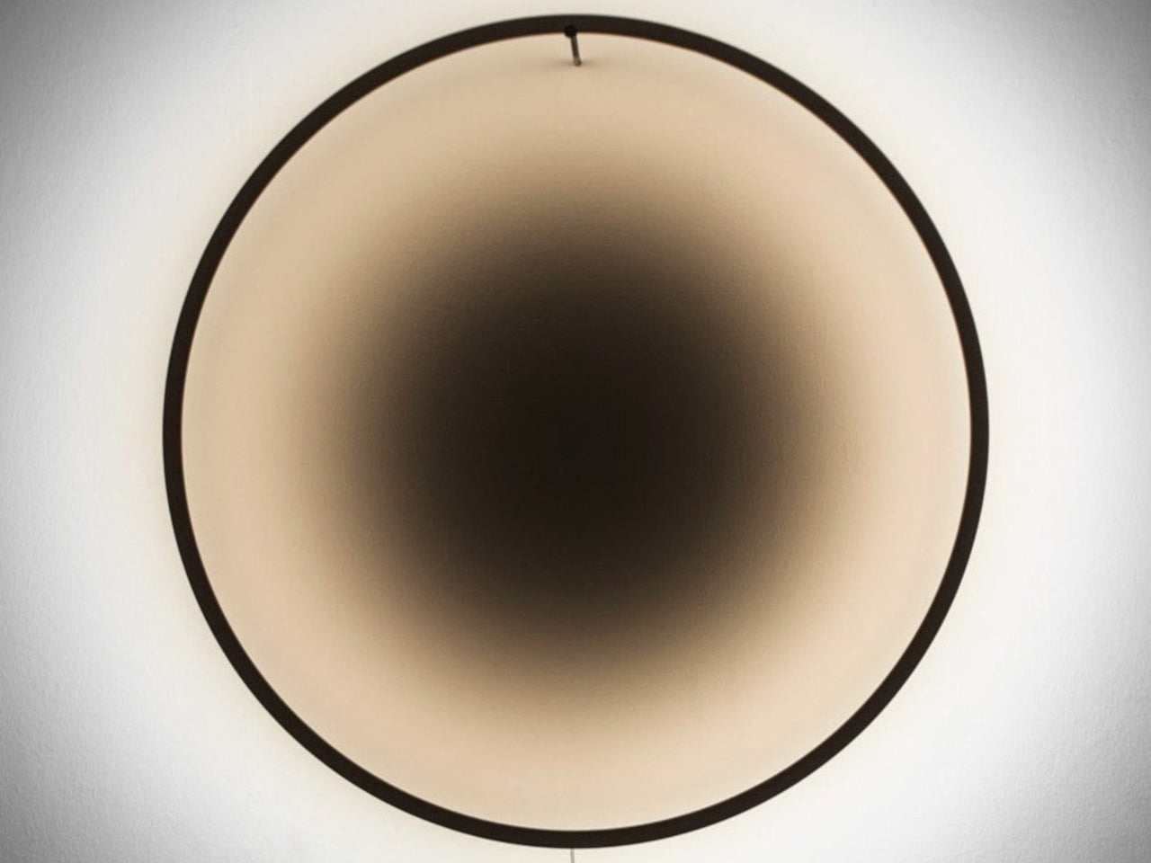 Round wall-mounted light fixture with a black rim and a warm beige gradient shade, creating a soft glow against a white wall.