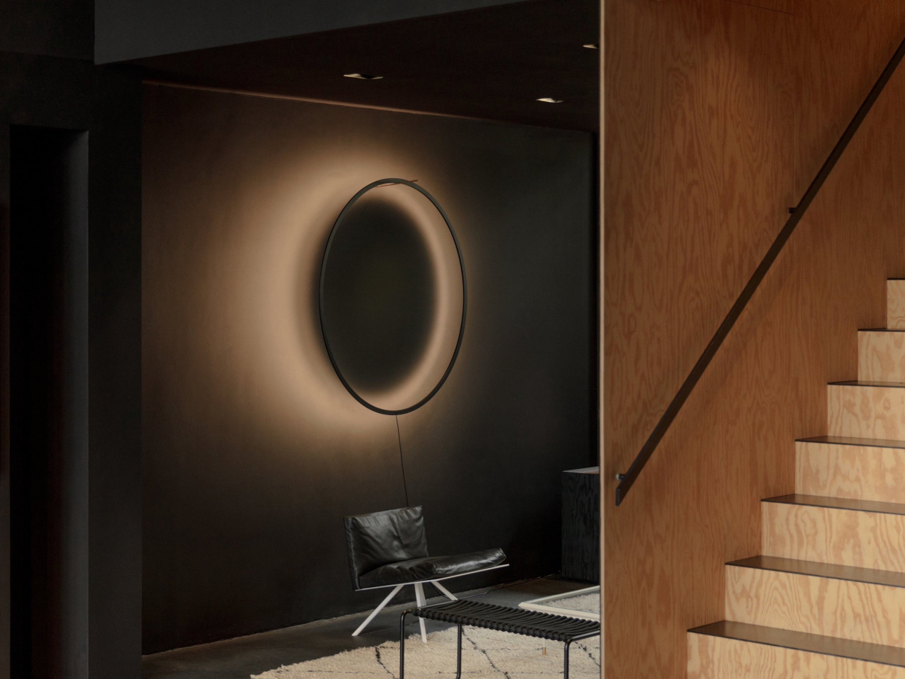Minimalist living area with a circular halo light on a dark wall, a black leather chair, and a wooden staircase to the right.