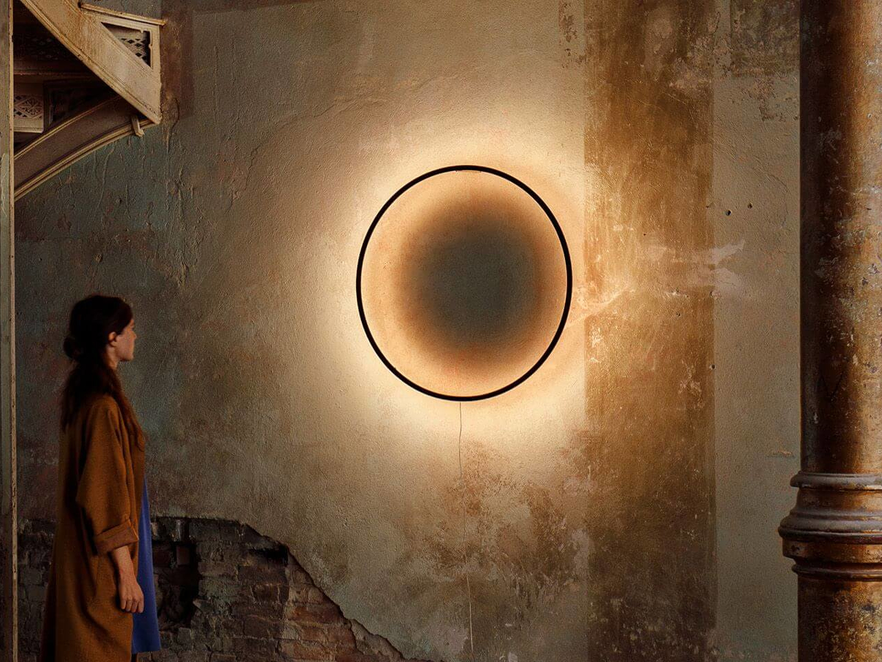 Woman in a brown coat standing in a weathered room, looking at a large glowing circular artwork on the wall.