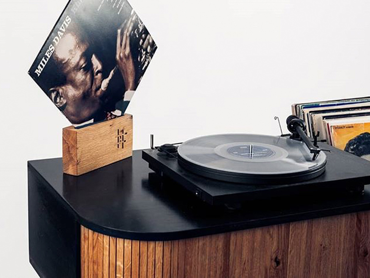 Turntable on a dark cabinet with a stack of vinyl records in the background and a Miles Davis album cover propped on a wooden stand.