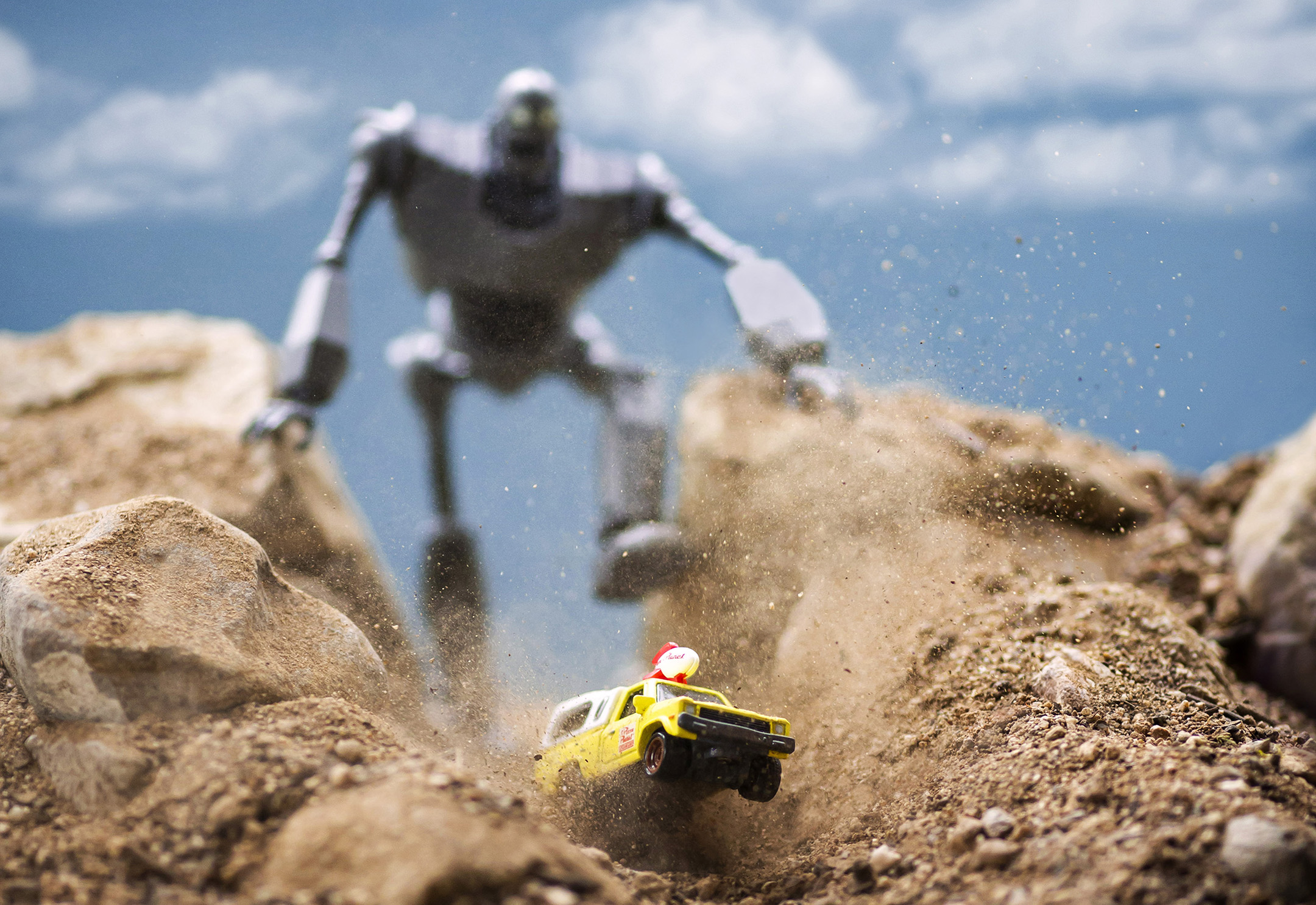 Toy yellow car kicking up dirt while fleeing a large, chased by blurred robot figure among rocks under a blue sky