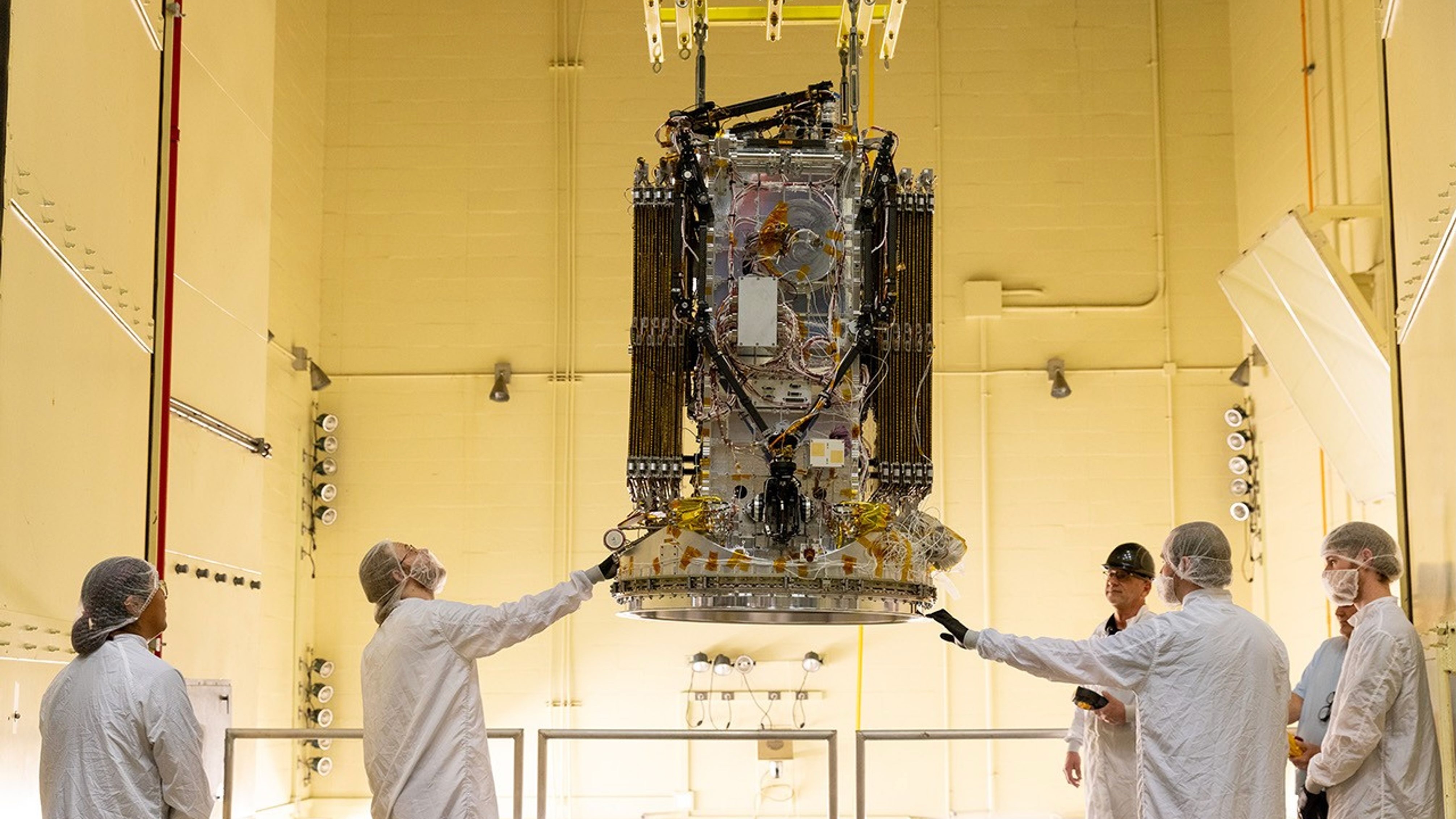 five people wearing white protective equipment stand around a rectangular spacecraft, which is hanging from the ceiling of a white-walled clean rooom