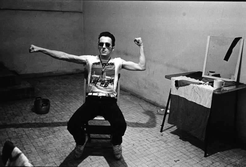A person wearing sunglasses and a “The Clash” t-shirt sits on a chair in a sparse room, flexing both arms. A table with a mirror, towel, cup, and kettle are next to them. The setting appears simple and industrial.