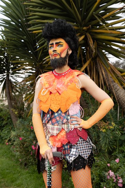 A person with dramatic, colorful makeup and a painted beard wears a patchwork outfit and orange fishnet gloves, standing in a garden with large spiky plants in the background.