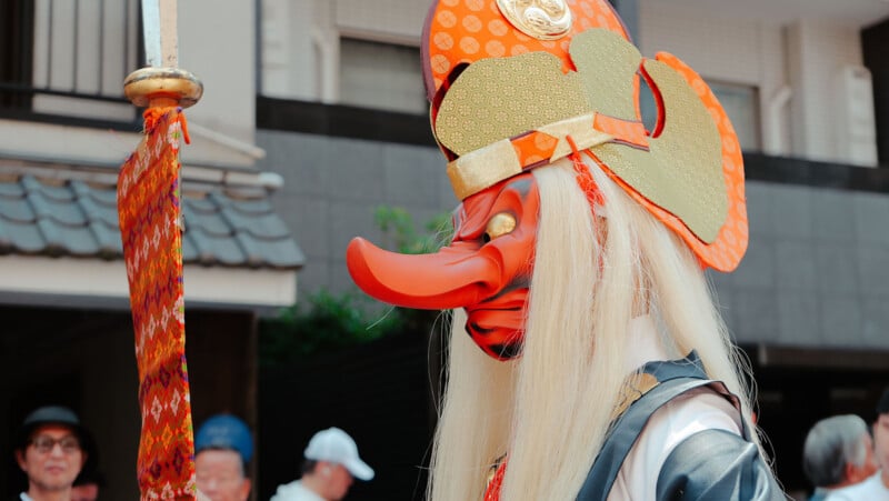 A person wearing a traditional Japanese Tengu costume with a red mask featuring a long nose, golden hat, and long white hair, standing outdoors during a festival with people in the background.