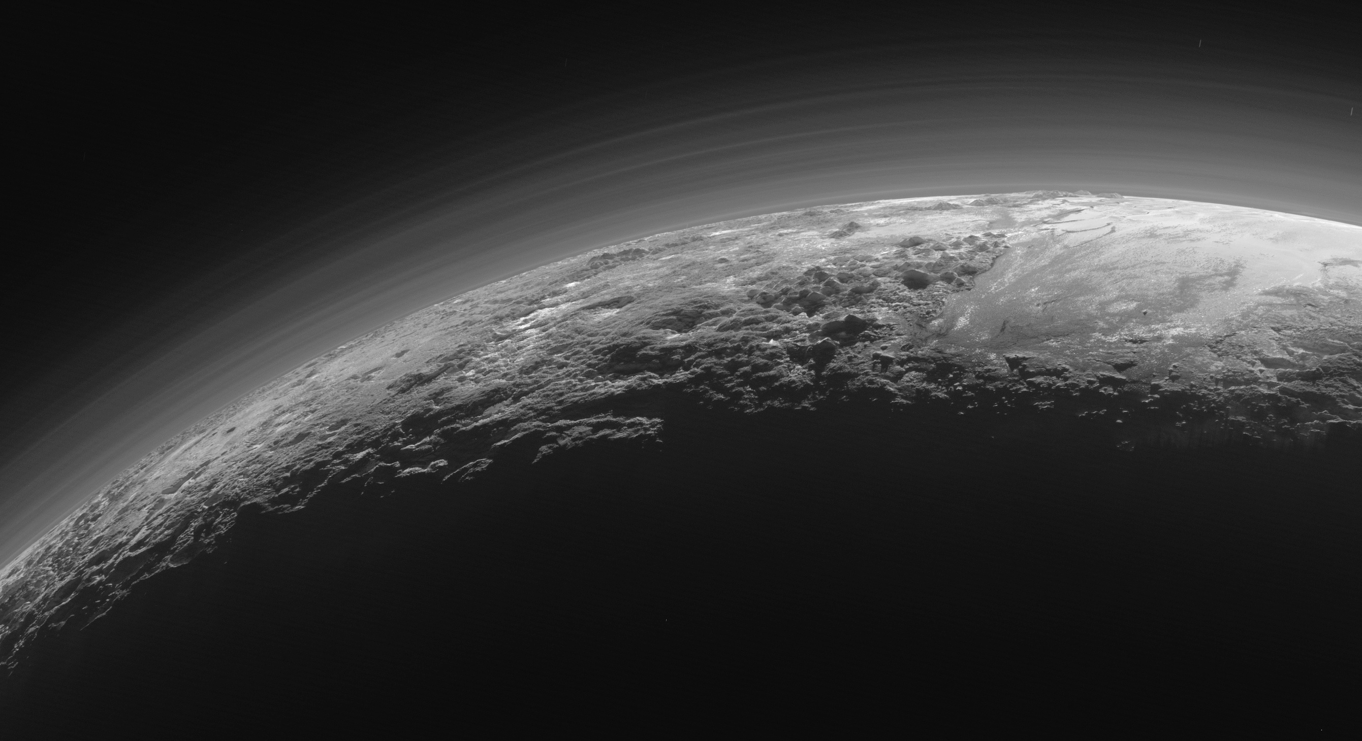 A black-and-white image shows the rugged surface and thin, hazy atmosphere of Pluto, a distant world in the Kuiper Belt, with the planet’s curvature visible against the dark background.