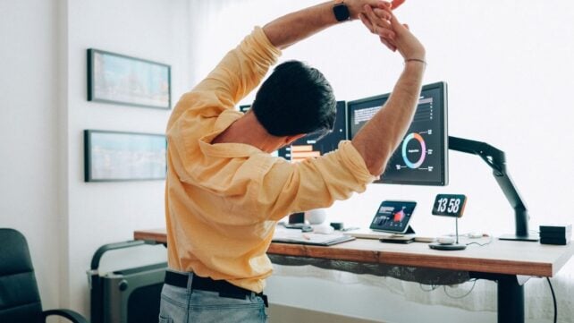 Brunette man in yellow shirt at work desk, stretching arms overhead