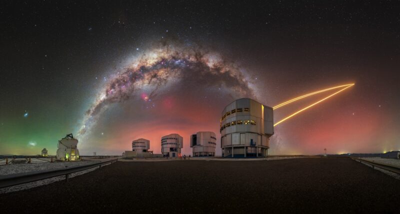 A row of telescope domes with a yellow laser on the right and the Milky Way arching overhead.