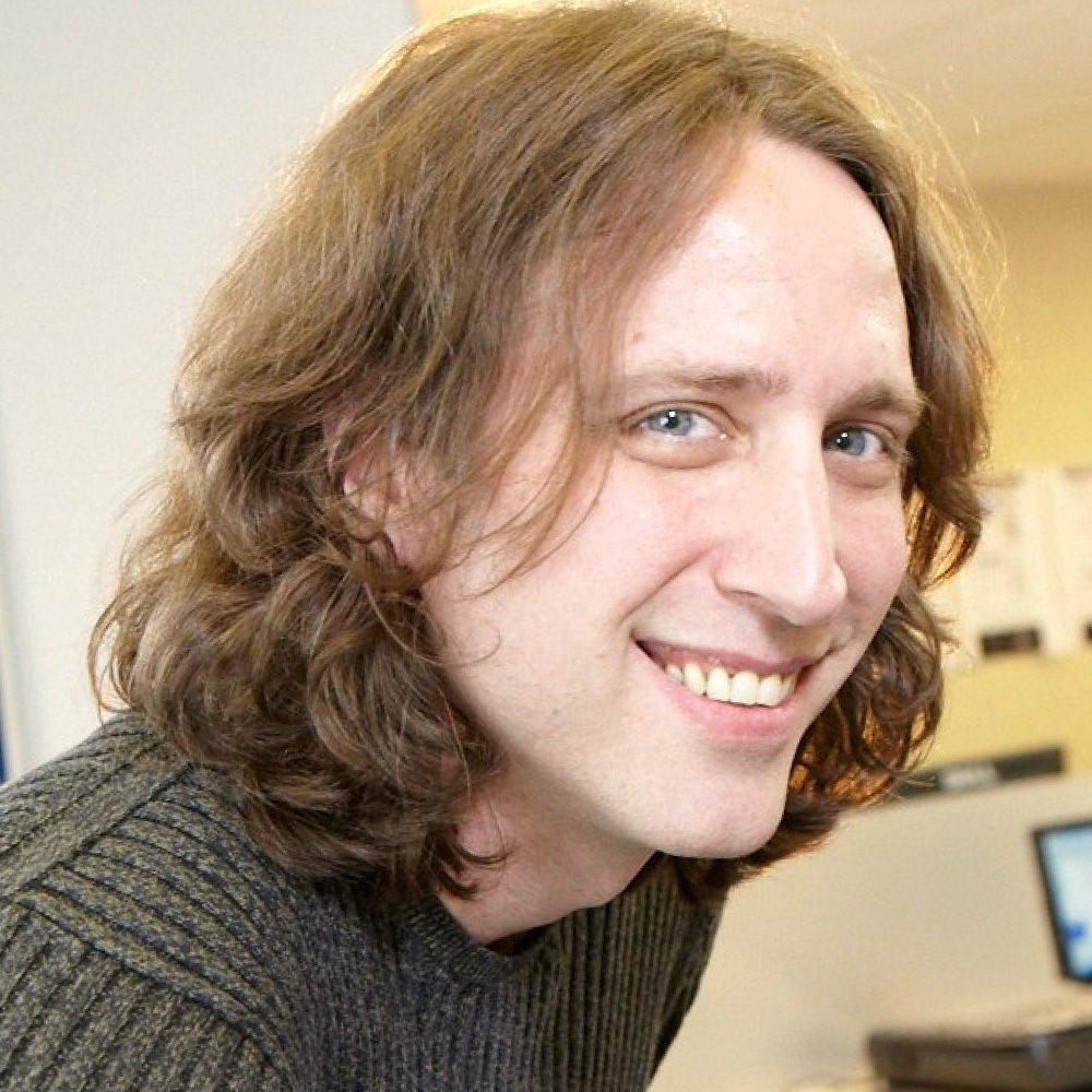 A photo of a smiling man in a jumper with long hair.
