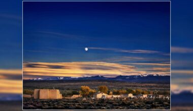 Adobe church and graveyard under a twilight sky with a full moon and snow-capped mountains in the distance.