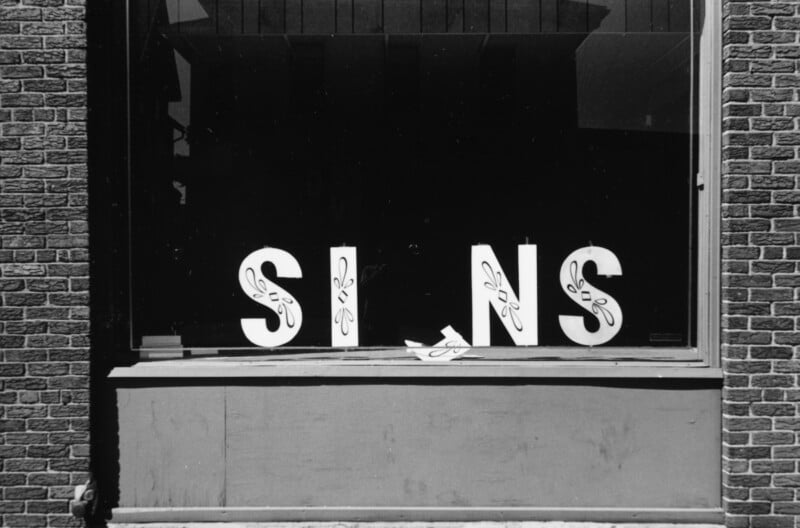 A storefront window displays large white letters spelling “SIGNS,” but the letter “G” has fallen, making it read “SINS.” The window is bordered by brick walls and has a dark interior.