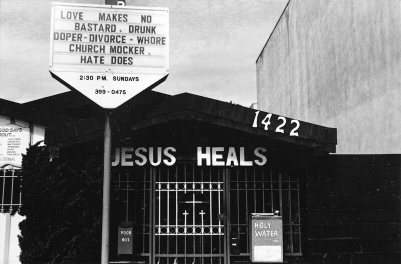 A black-and-white photo of a church with a sign reading “LOVE MAKES NO BASTARD. DRUNK. DOPER-DIVORCE-WHORE. CHURCH MOCKER. HATE DOES.” The building says "JESUS HEALS" above a gated entrance.