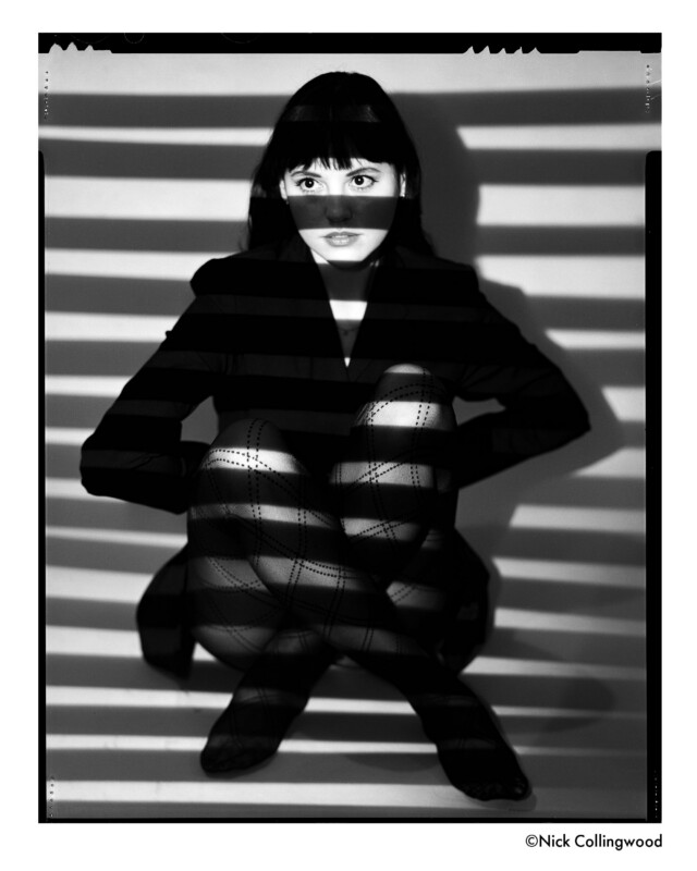 A woman sits with her knees up, wearing patterned tights and a dark outfit. Shadows from horizontal blinds fall across her face and body, creating a dramatic, striped effect in black and white.