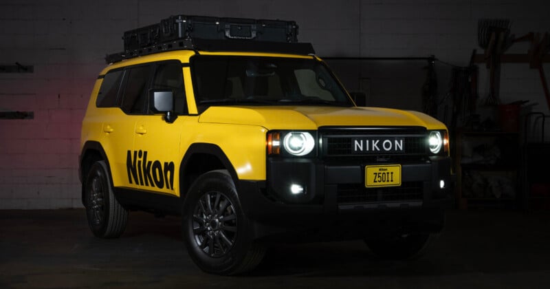 A yellow SUV with “Nikon” branding on the side and front, parked indoors. The vehicle has a roof storage box, black wheels, and a custom license plate reading “Z50II.” The headlights and fog lights are on.
