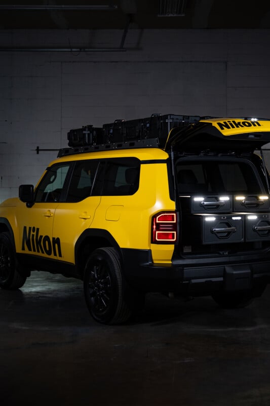 A yellow SUV with "Nikon" branding on the side and rear is parked indoors with its rear hatch open, revealing organized storage cases inside. The vehicle has black wheels and roof racks.