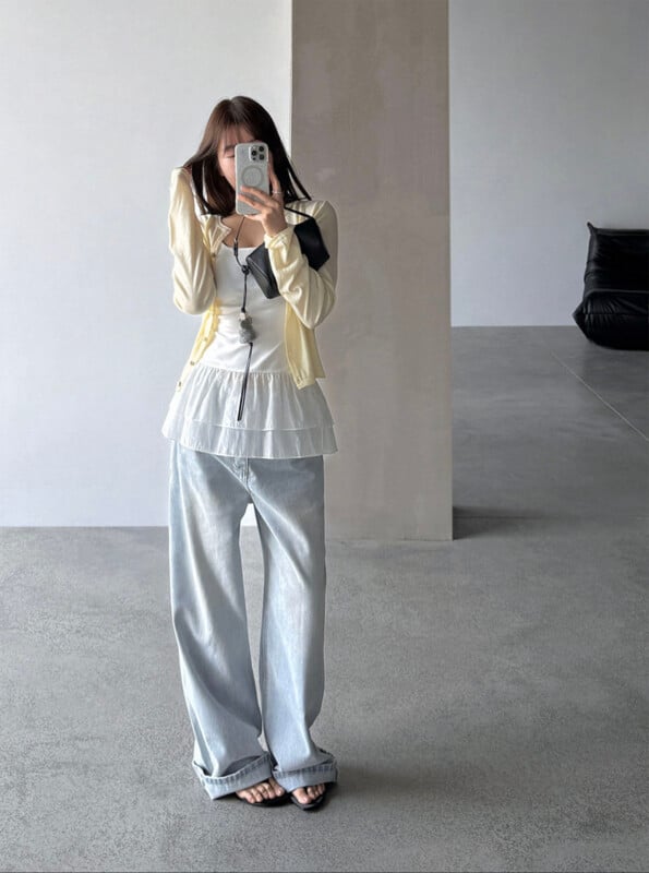 A woman stands indoors taking a mirror selfie. She wears a light yellow cardigan, a white layered top, loose light blue jeans, and black sandals. The minimalist room has gray floors and a black couch in the background.
