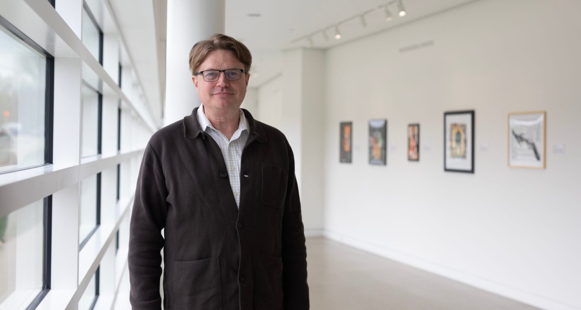 A man wearing glasses and a dark jacket stands in a bright art gallery hallway with framed artworks on the white wall behind him.