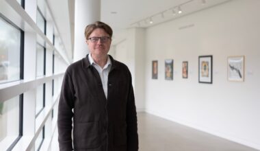 A man wearing glasses and a dark jacket stands in a bright art gallery hallway with framed artworks on the white wall behind him.