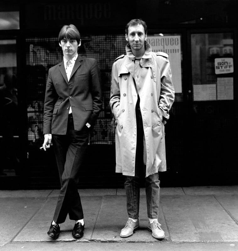 Two men stand on a city sidewalk. One wears a dark suit and holds a cigarette, while the other wears a trench coat, jeans, and sneakers. They pose in front of a building with "marquee" signage in the background. Black and white photo.