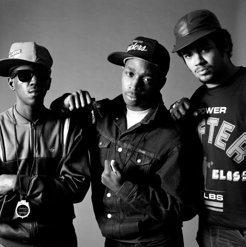 Three men pose together in black and white, wearing baseball caps and streetwear. One wears sunglasses and a stopwatch, the middle man is in denim, and the third wears a sweatshirt with “LIFTER” printed on it.