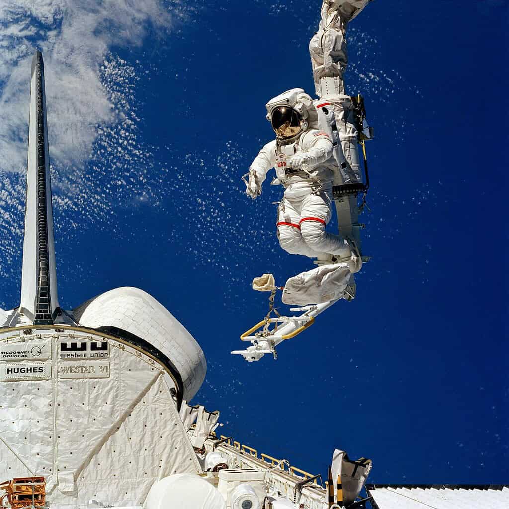Astronaut performing a spacewalk outside the ISS in a white suit against a blue sky.