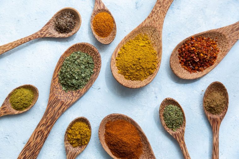 Flat lay arrangement of dried ground pungent spices and aromatic herbs in rustic wooden spoons in a random scatter on a blue background viewed from above