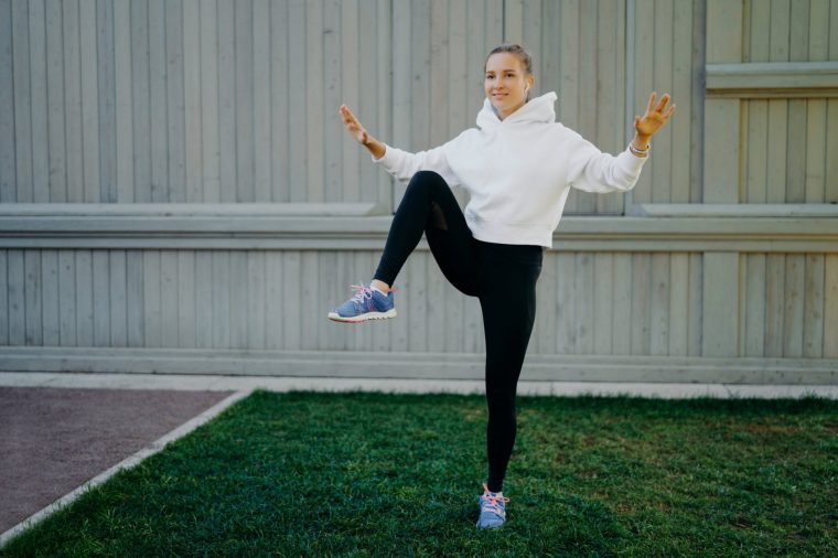 Energetic sportwoman has workout routine poses on one legs demonstrates her stamina warms up before jogging dressed in hoodie leggings and trainers trains outdoors. Sport and motivation concept