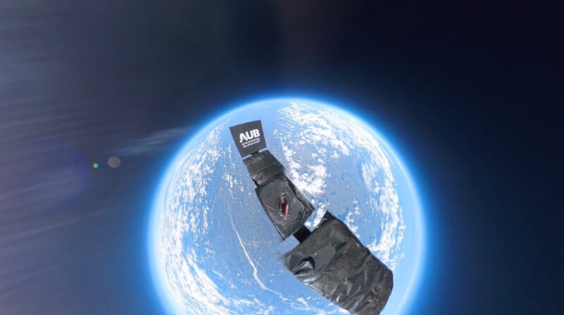 A small satellite or CubeSat, with "AUB" printed on it, orbits above Earth against the blackness of space, with the planet’s blue and white surface visible below.