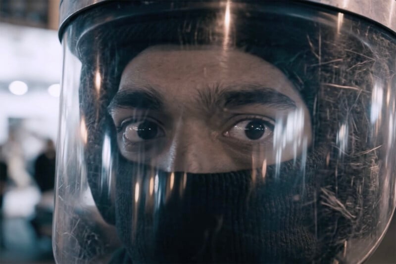 Close-up of a person's face wearing a protective helmet with a clear visor and a black balaclava, showing only their eyes, looking alert and intense. The background is blurred.