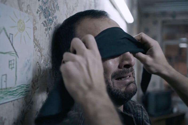 A man with a distressed expression is being blindfolded with a black cloth by another person's hands. He appears to be inside a modest room with patterned wallpaper and a child's drawing on the wall.