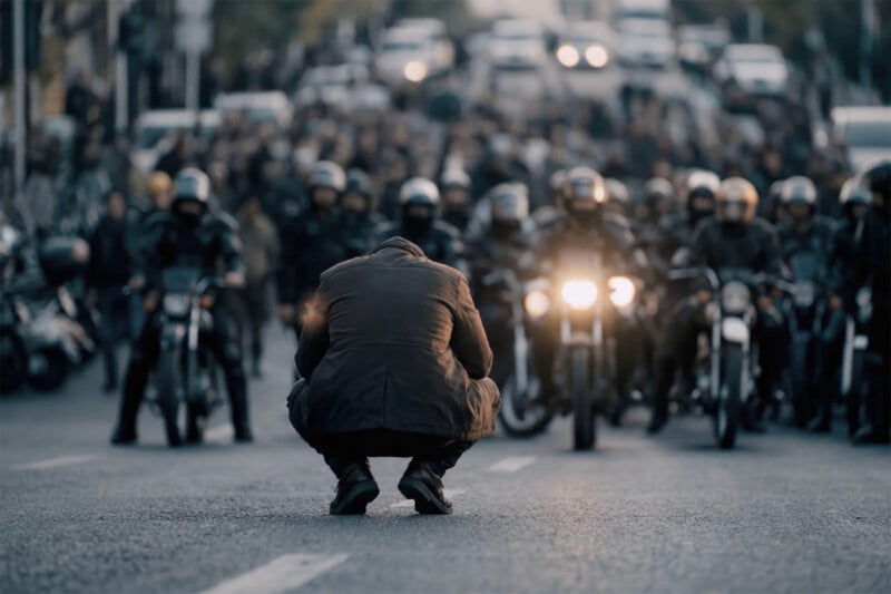 A person crouches alone in the middle of a street facing a group of motorcyclists and a large crowd behind them, creating a tense and dramatic atmosphere.