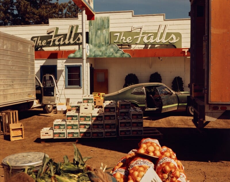 A stack of produce boxes and bags of onions sits outside “The Falls” storefront. A green car with an open door is parked nearby, surrounded by trucks. The scene appears to be a market or delivery area.