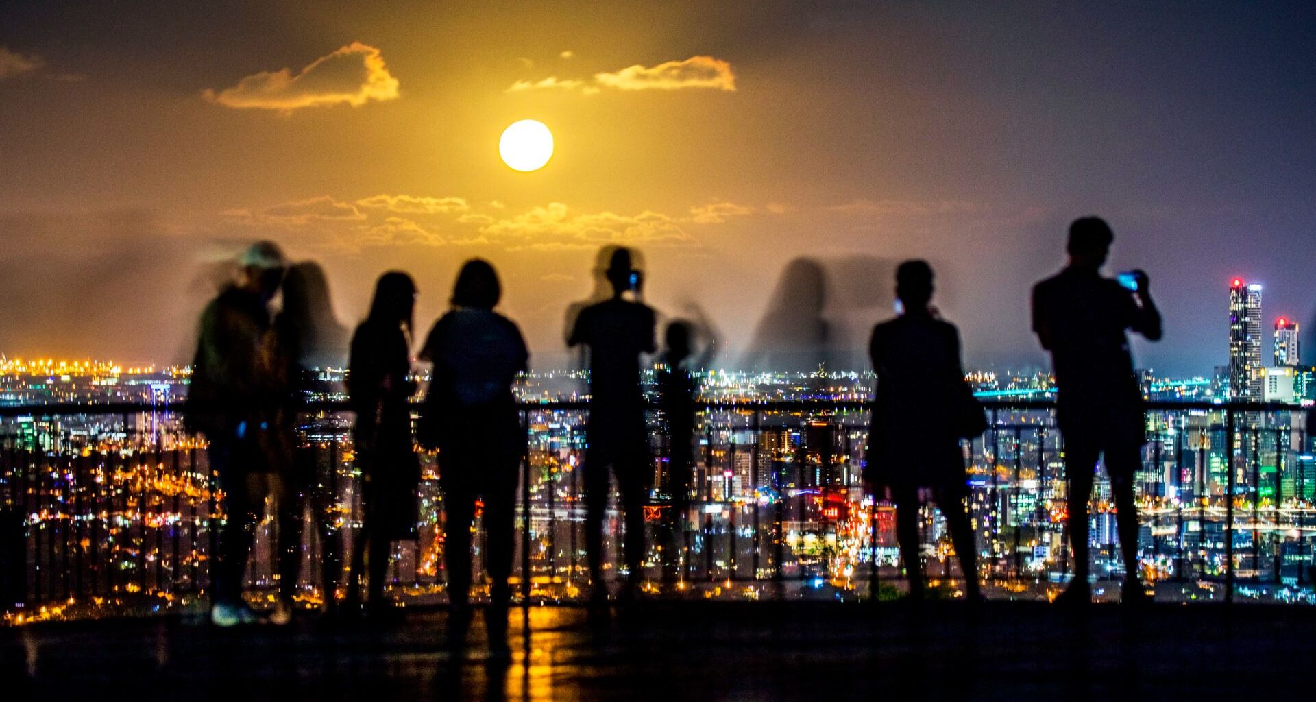 Silhouettes of people standing on a balcony overlook the cityscape of lights below with a full glowing moon in the distance.