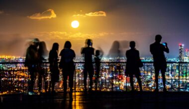 Silhouettes of people standing on a balcony overlook the cityscape of lights below with a full glowing moon in the distance.