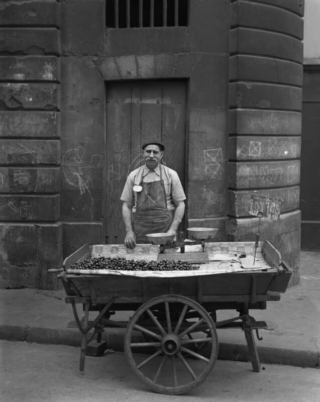 A man wearing an apron and hat stands behind a wooden pushcart filled with produce, possibly olives, on a city street. He is positioned in front of a weathered stone building with chalk drawings on the wall.