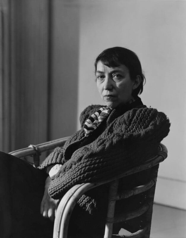 A woman with dark hair wearing a textured, chunky knit sweater and patterned scarf sits in a wicker chair, looking directly at the camera with a serious expression in a softly lit room.