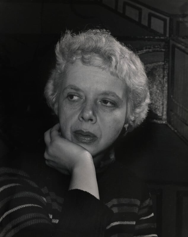A person with short, curly light hair rests their chin on their hand, looking thoughtfully to the side. They wear a striped sweater. The background is dark with geometric shapes visible.