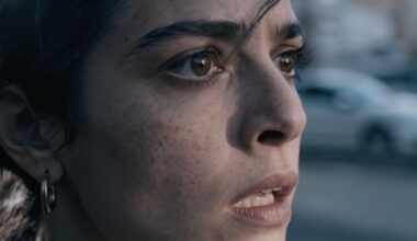 A close-up of a woman with dark hair and brown eyes, looking ahead with a worried or anxious expression. Her mouth is slightly open, and the background appears blurred, showing an outdoor scene.